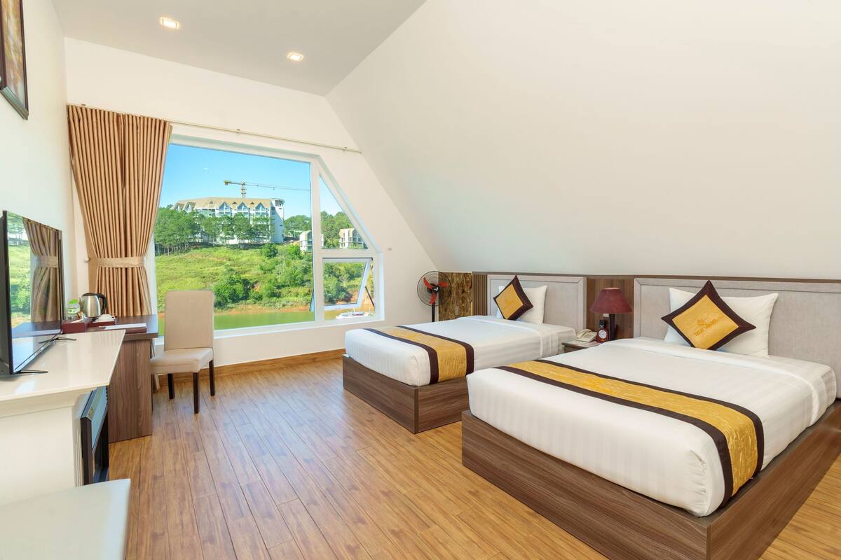 Ein Hotelzimmer mit zwei Betten, einem Schreibtisch, einem Sessel und einem großen Fenster mit Blick auf Grünflächen und Gebäude.