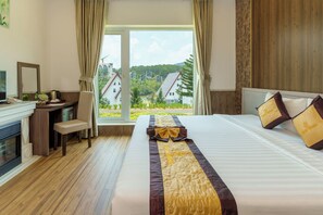Une chambre d’hôtel avec un lit, un bureau, une chaise, une cheminée et une grande fenêtre donnant sur des maisons et des arbres.