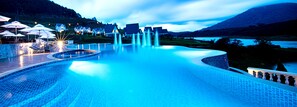 Une piscine à débordement offrant une vue sur un lac et des montagnes, entourée de parasols et de chaises longues.