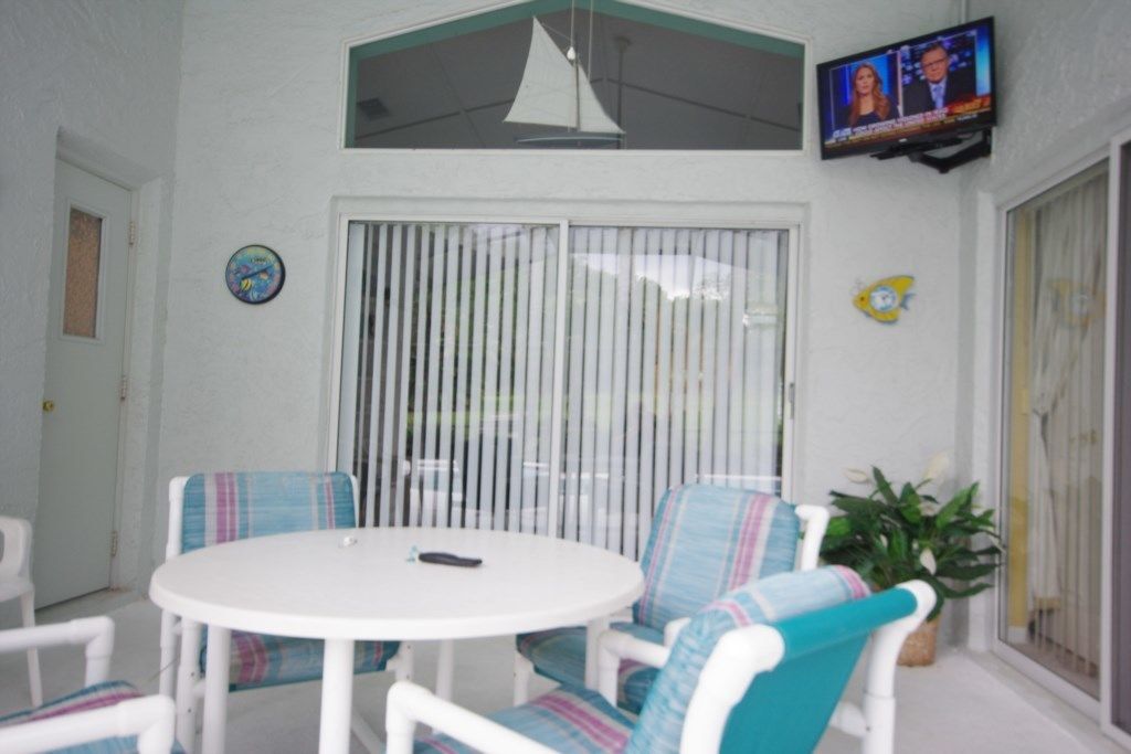 A room with a round table and chairs, a television mounted on the wall, and a window with vertical blinds.