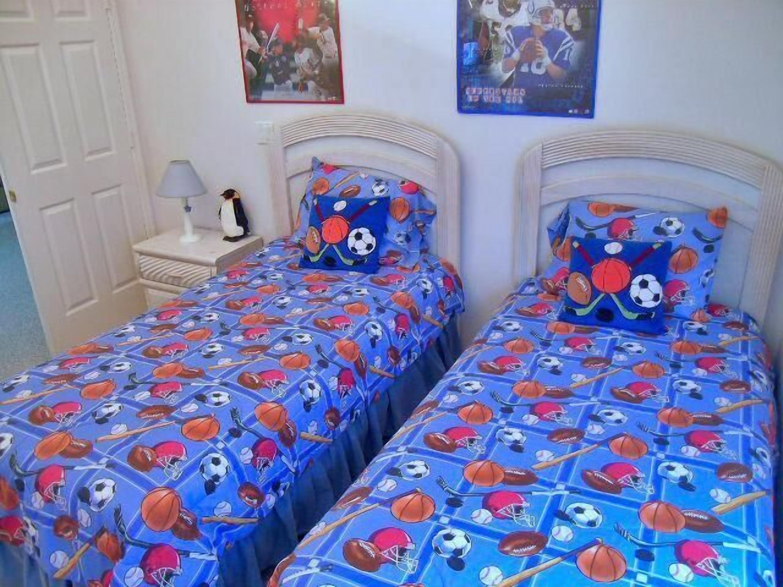 A bedroom with two beds, each with a blue bedspread featuring sports equipment. There is a nightstand with a lamp and a small decorative item on it.