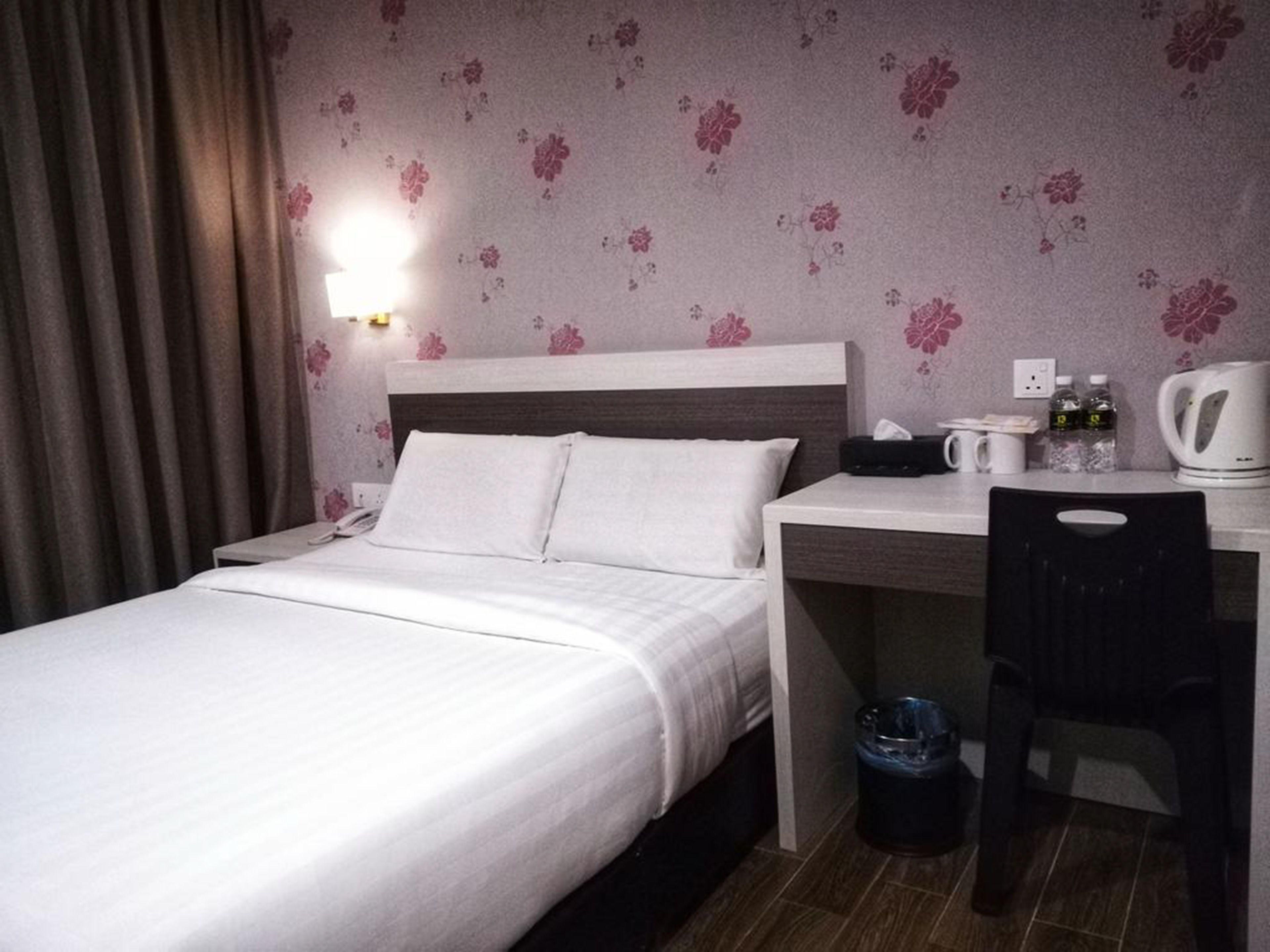 Habitación de hotel con cama, escritorio, silla, cesto de basura, cafetera y papel tapiz floral en la pared.