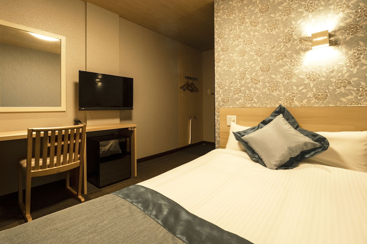 A hotel room with a bed, a desk, a chair, a television, and a wall-mounted light.