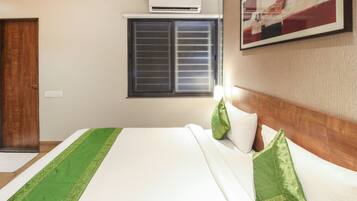 A hotel room with a bed, a window with blinds, an air conditioning unit, and a painting on the wall.