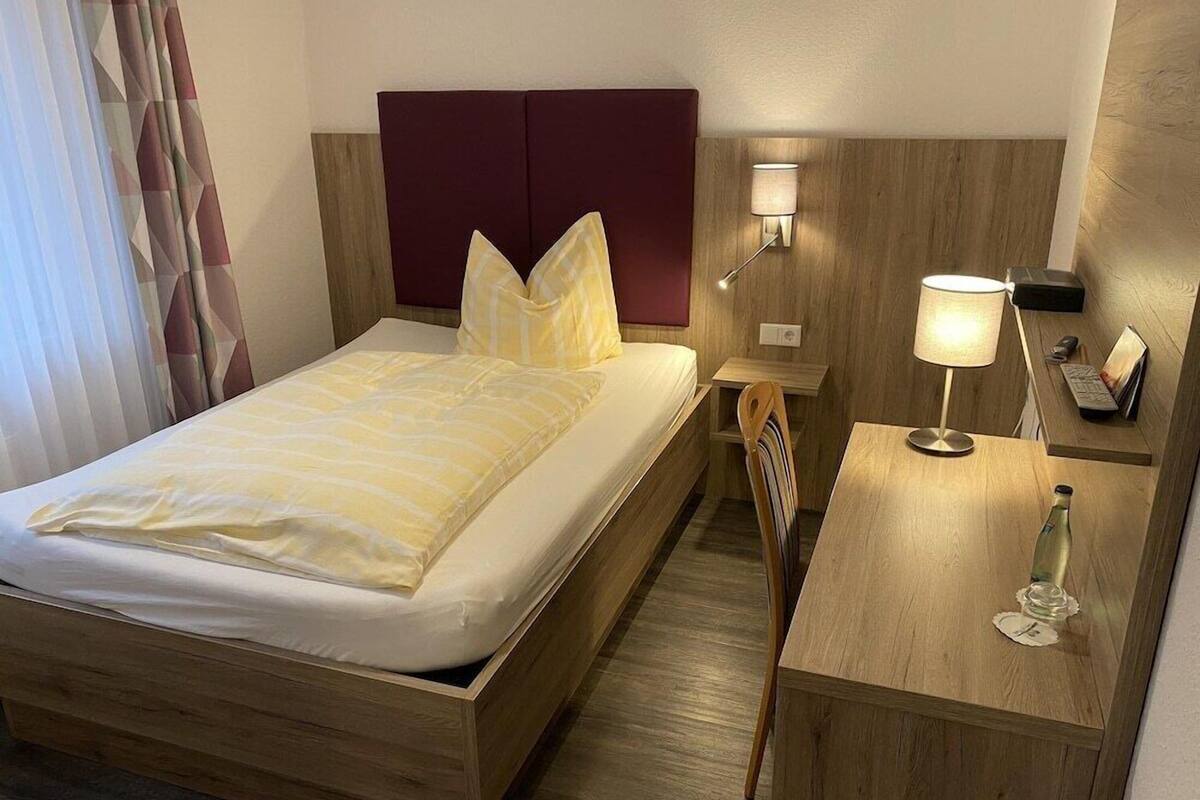 Une chambre d’hôtel avec un lit en bois, un bureau avec une lampe, une chaise et une table de chevet avec un téléphone et une bouteille.