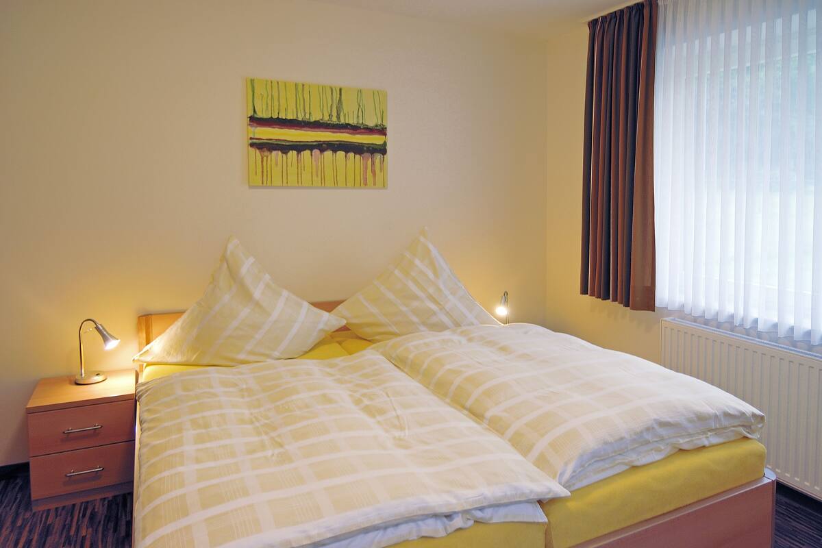 Un lit double avec une couvre-lit à carreaux jaunes et blancs, une lampe de chevet et une table de chevet avec des tiroirs.