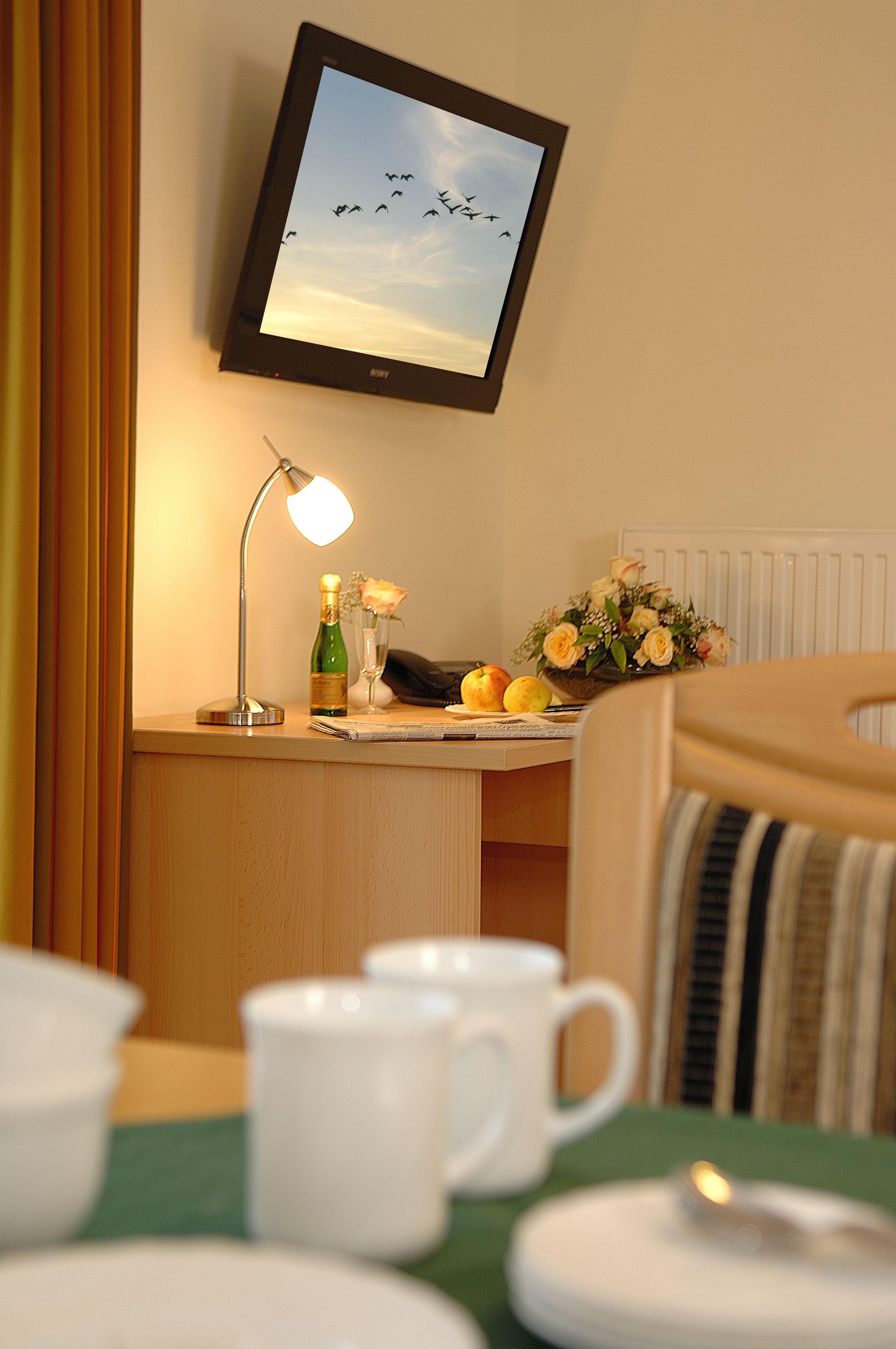 Habitación de hotel con televisión mostrando aves volando, una mesita de noche con lámpara, un florero con flores y un frutero.