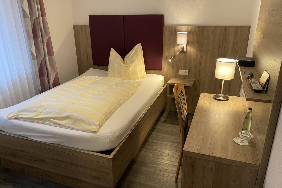 Un lit bien fait, avec une tête de lit, une table de chevet avec une lampe, une chaise et un petit bureau sur lequel se trouvent une bouteille et des verres.