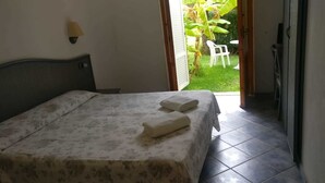 A hotel room with a bed, a nightstand, a wall-mounted lamp, and a view of a garden with a chair outside. - Hotel Club Torre Marino (Ricadi)
