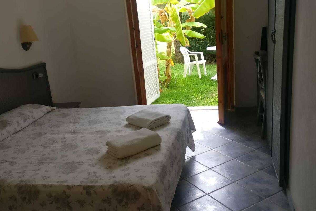 A hotel room with a bed, a nightstand, a wall-mounted lamp, and a view of a garden with a chair outside.