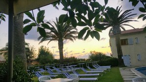 A poolside area with lounge chairs and tables, surrounded by palm trees and a building with a tiled roof. - Hotel Club Torre Marino (Ricadi)