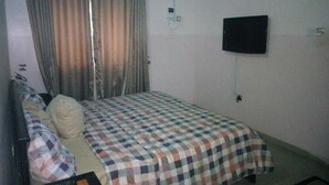 A single bed with a checkered bedspread, a television mounted on the wall, and a window with curtains. - Bayse One Executive Suites and Bar (Ibadan)