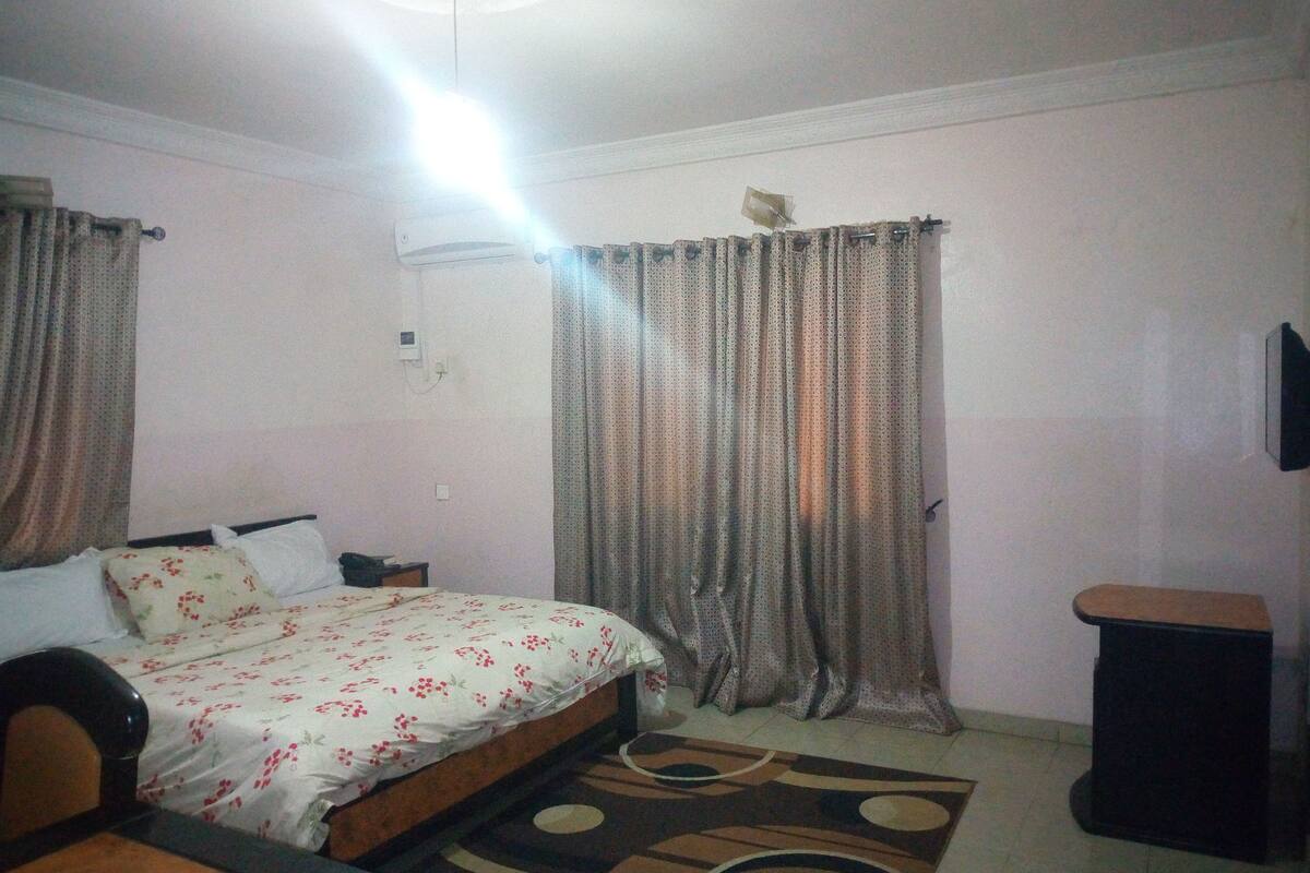 A bedroom with a bed, curtains, a small table, a chair, a rug, and a wall-mounted TV.
