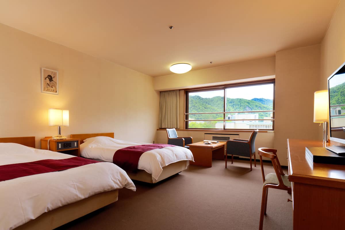 A hotel room with two beds, a desk, a chair, a TV, and a window with a view of greenery.