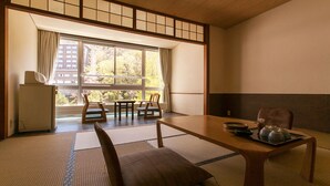 Una habitación tradicional japonesa con suelo de tatami, una mesa baja con una bandeja de utensilios de té y vistas a un edificio a través de un gran ventanal.