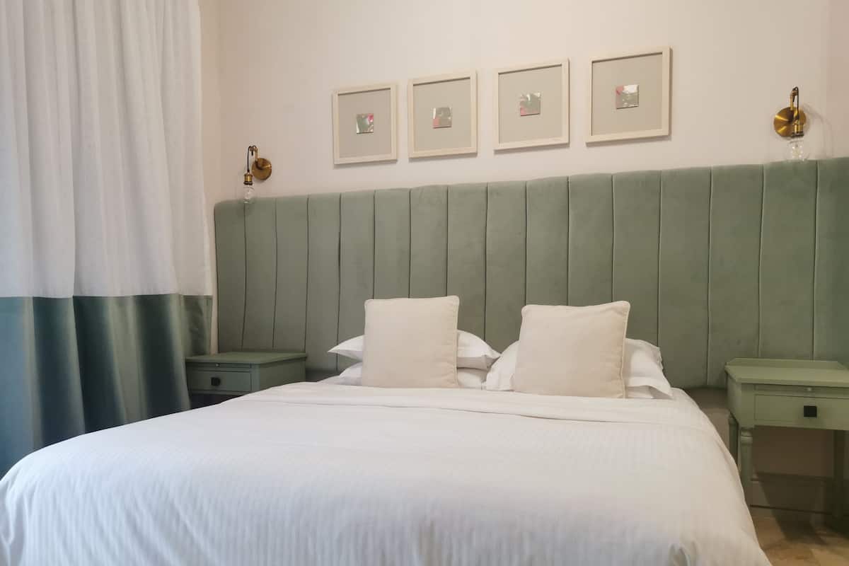 Una cama bien hecha con sábanas blancas, un cabecero tapizado en verde y una mesita de noche con una lámpara.