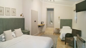 Habitació d'hotel amb un llit gran, un llit més petit, una taula, una cadira, un mirall, una escala i quadres amb marc a la paret.