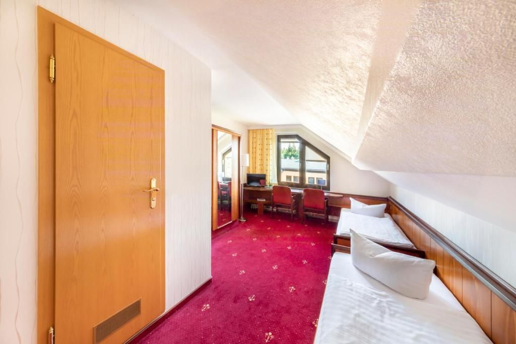 Una habitación con puerta de madera, alfombra roja, cama con almohadas, escritorio con sillas y ventana con vistas.
