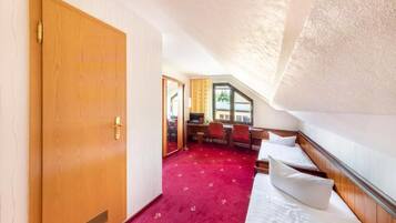 Una habitación con puerta de madera, alfombra roja, cama con almohadas, escritorio con sillas y ventana con vistas.