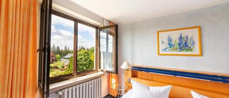 A hotel room with a bed, a window with curtains, a radiator, a lamp, and a framed picture on the wall.