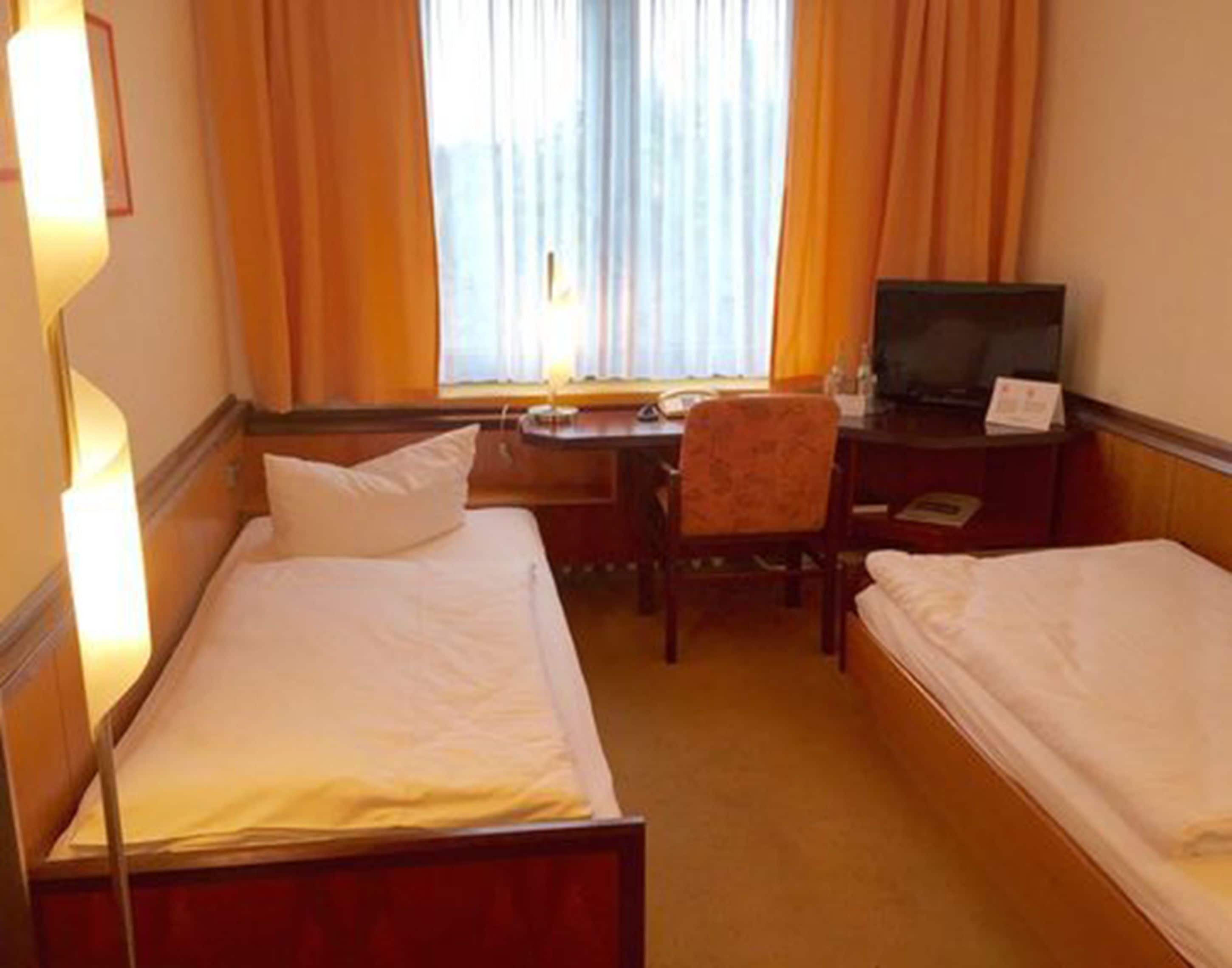 Habitación de hotel con dos camas individuales, un escritorio con silla, televisor y ventana con cortinas.