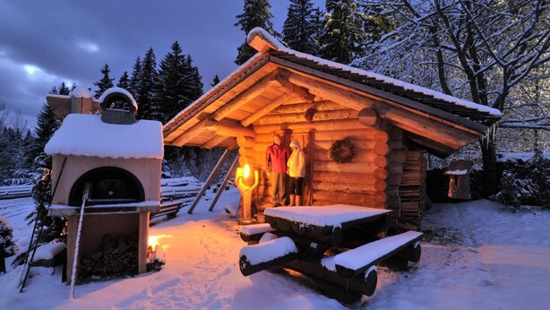 Una cabaña de troncos con chimenea, un banco y un terreno cubierto de nieve.