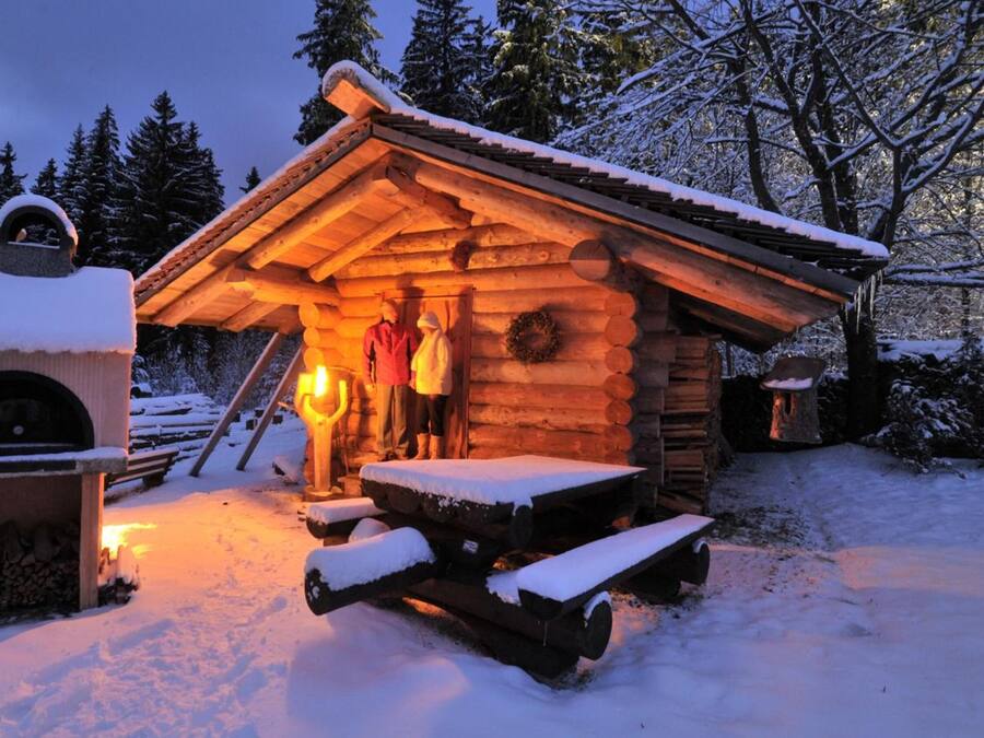 A log cabin with a fireplace, a bench, and a snow-covered ground.