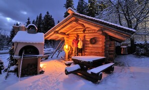 Una cabaña de troncos con chimenea, un banco y un terreno cubierto de nieve.