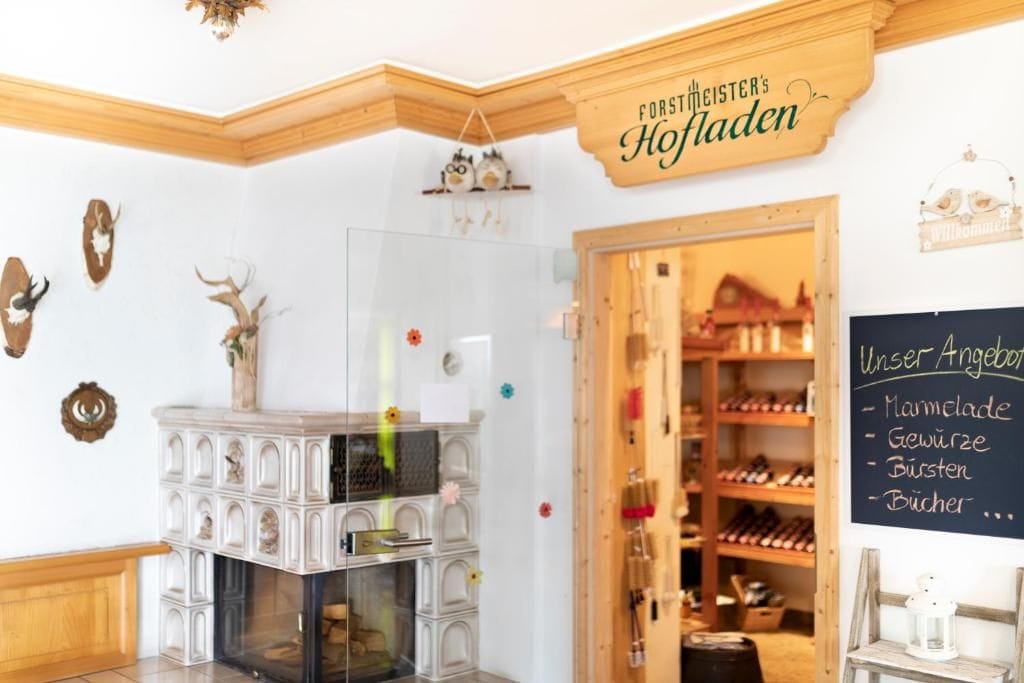 Un hall d’hôtel doté d’une cheminée, une enseigne indiquant « Forstmeister’s Hofladen », et un tableau noir affichant le menu avec des articles tels que de la confiture et des épices.