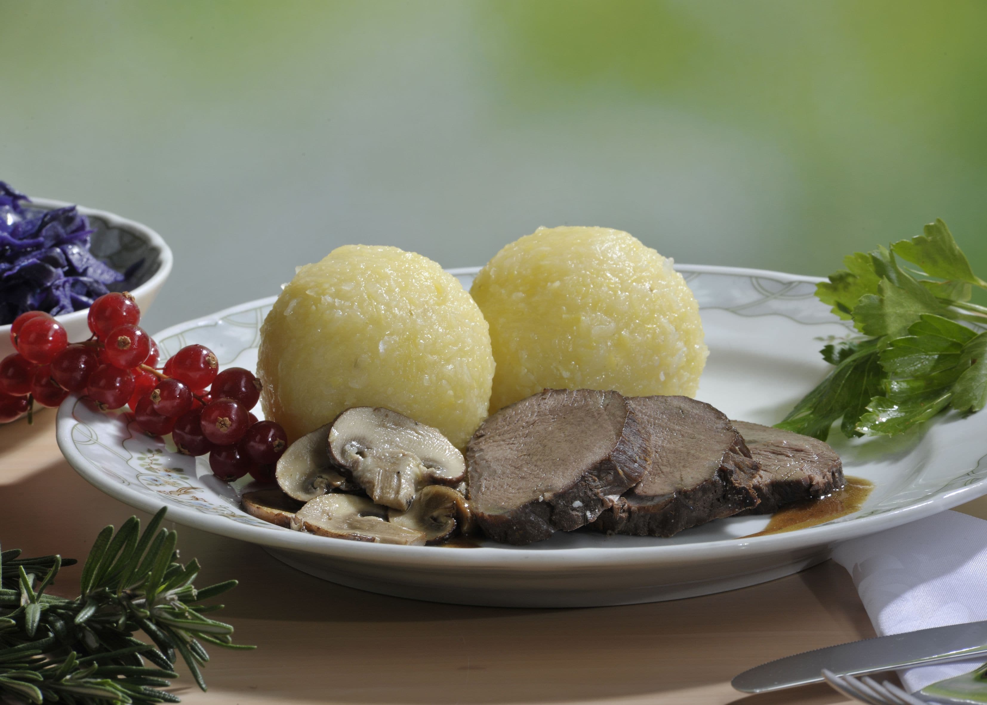 A plate with two round yellow dumplings, sliced beef, mushrooms, red currants, and a sprig of rosemary.