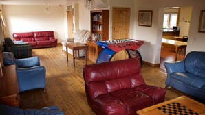 A living room with a red leather sofa, blue armchairs, a wooden coffee table with a chessboard, a foosball table, and a bookshelf. - Gilfach Wen Barn (Carmarthen)