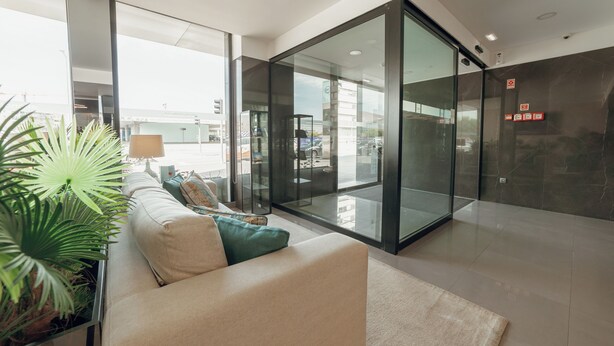 A modern hotel lobby with a glass partition, a sofa with cushions, a lamp, and a potted plant.