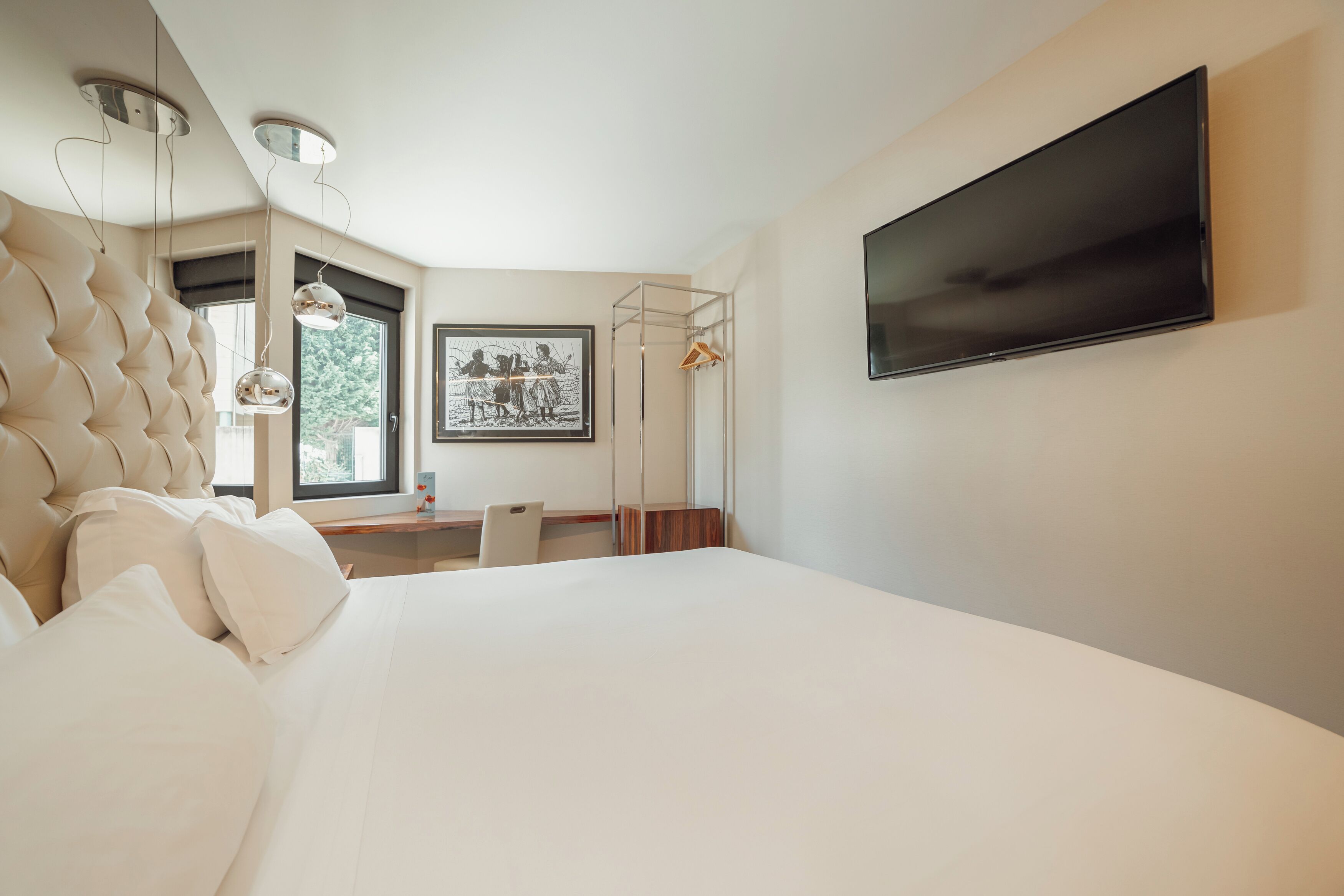 Un dormitorio moderno con una cama grande, un televisor montado en la pared, un escritorio con silla y una ventana con vistas a zonas verdes.