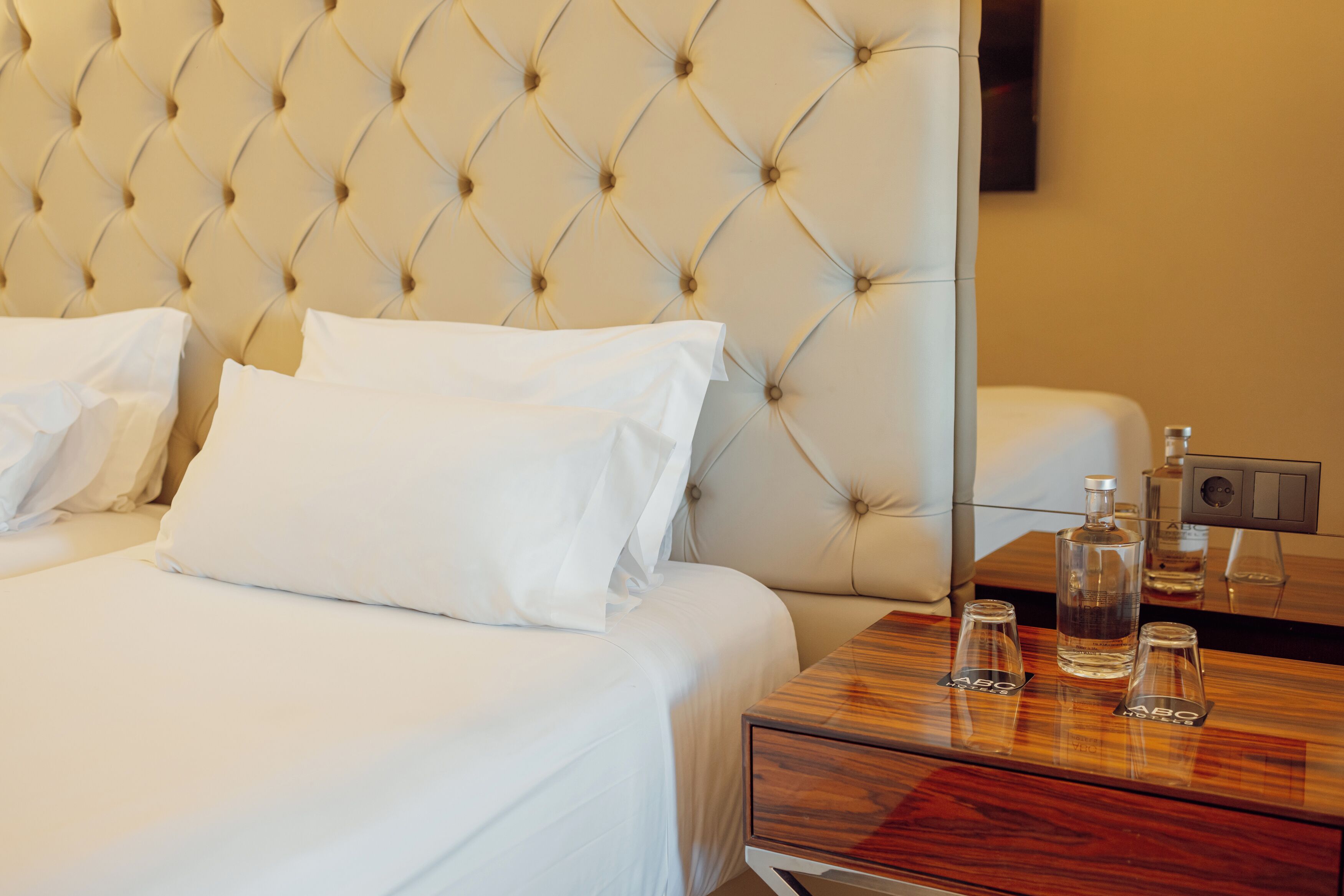 Une chambre d’hôtel avec une tête de lit capitonnée, du linge de lit blanc et une table de chevet en bois sur laquelle sont posées trois bouteilles et des dessous de verre.