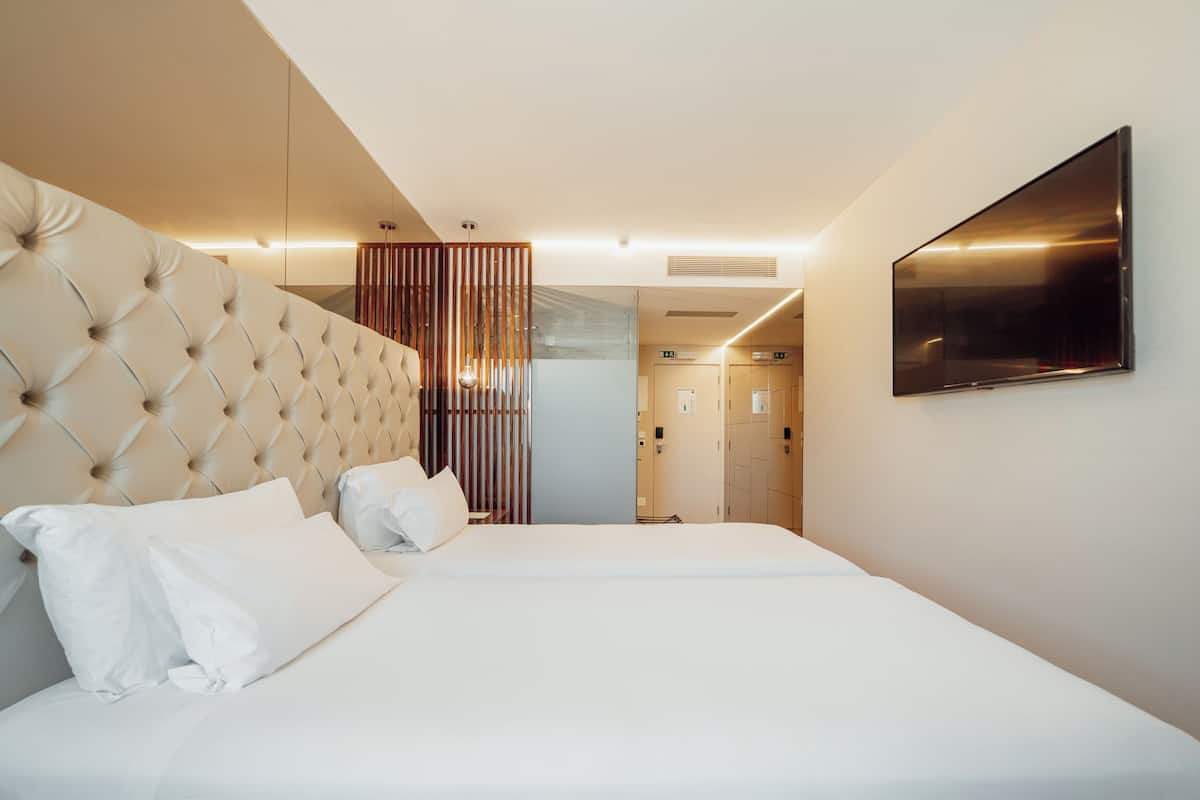 Ett modernt hotellrum med en stoppad sänggavel, en platt-TV som är monterad på väggen och en separat säng med vita sängkläder.