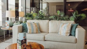 A modern hotel lobby with a beige sofa, decorative pillows, a round wooden coffee table, and a glass partition with potted plants.