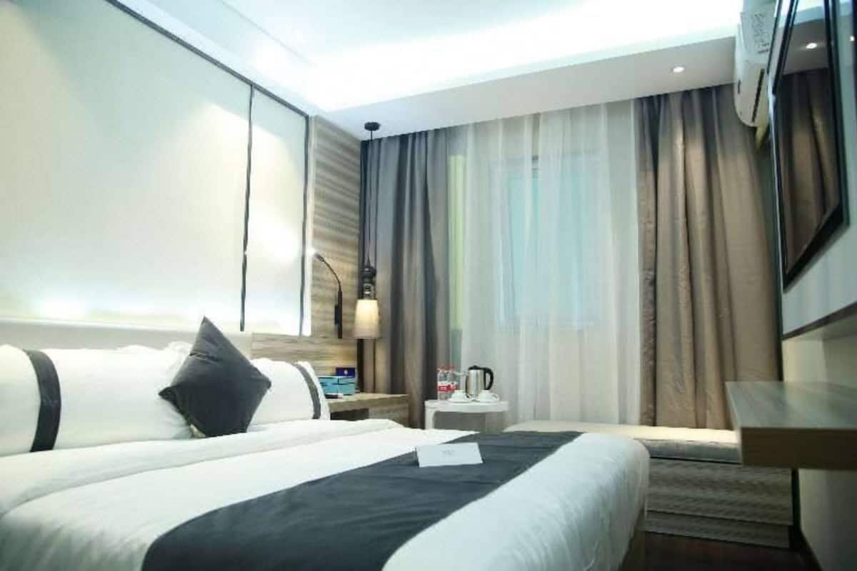 A modern hotel room with a large bed, a bedside table with a lamp, a small table with a coffee maker, and a large window with curtains.