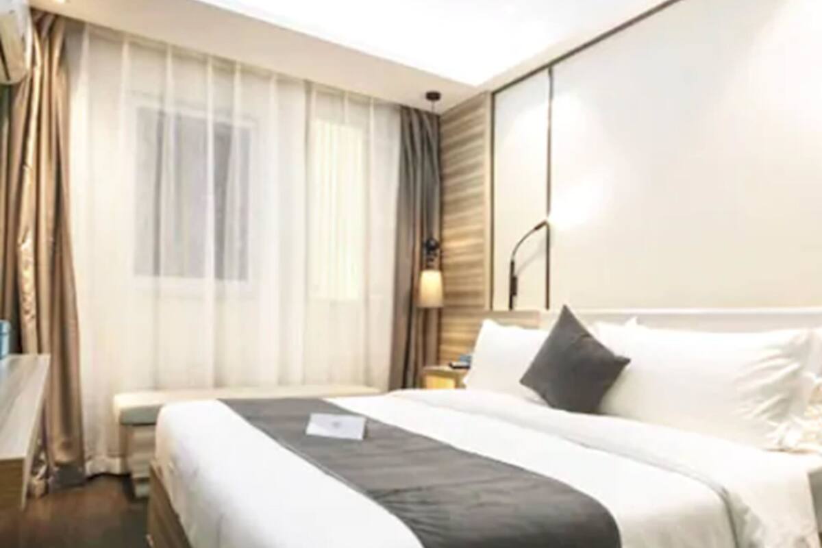 A hotel room with a large bed, a bedside table, a lamp, and a window with sheer curtains.