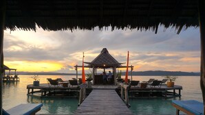 A wooden pier leading to a thatched-roof structure with lounge chairs and potted plants. - Raja Ampat Dive Lodge (Mansuar Island)
