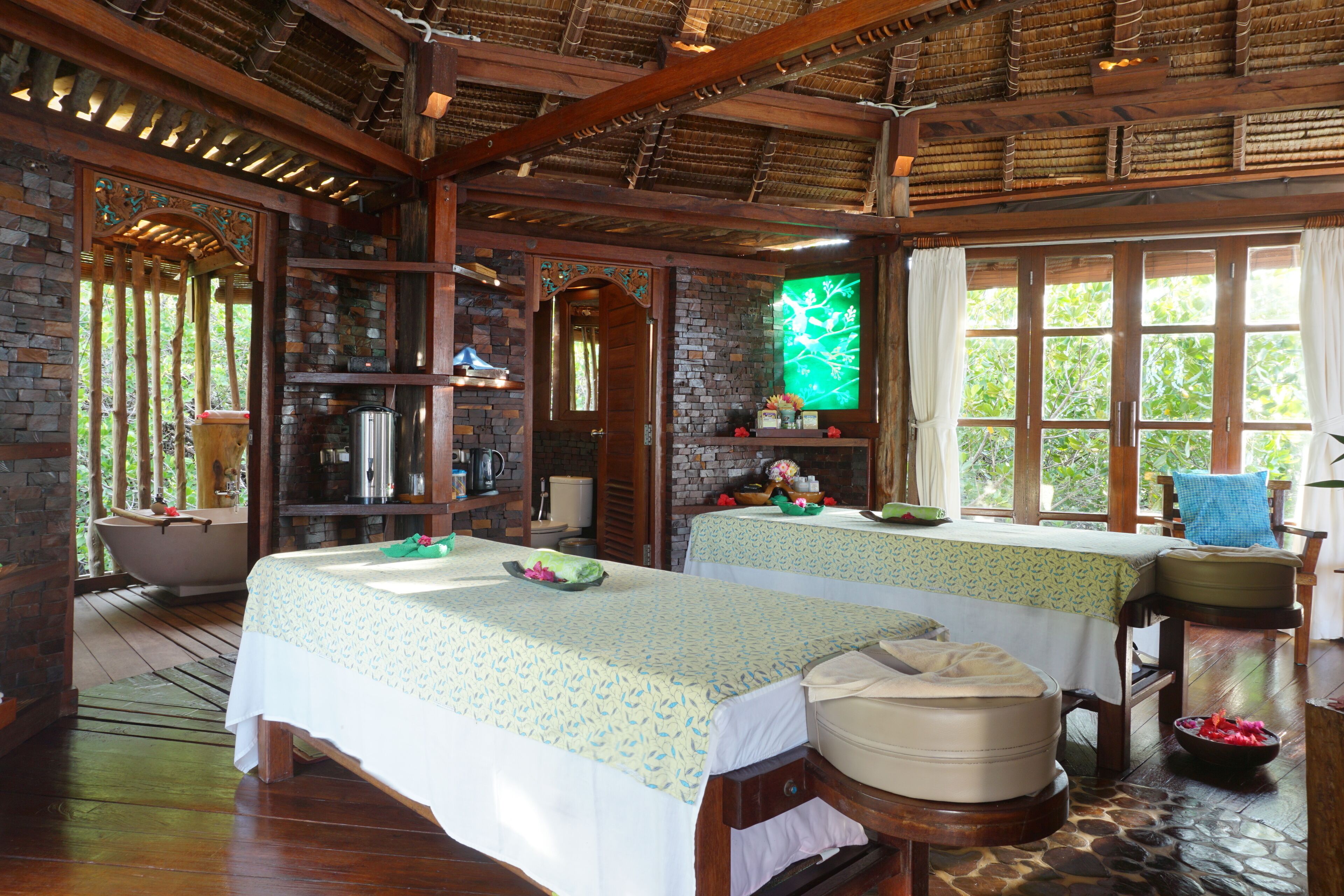 A room with two beds, a bathtub, a wooden ceiling, and a window with a view of trees.