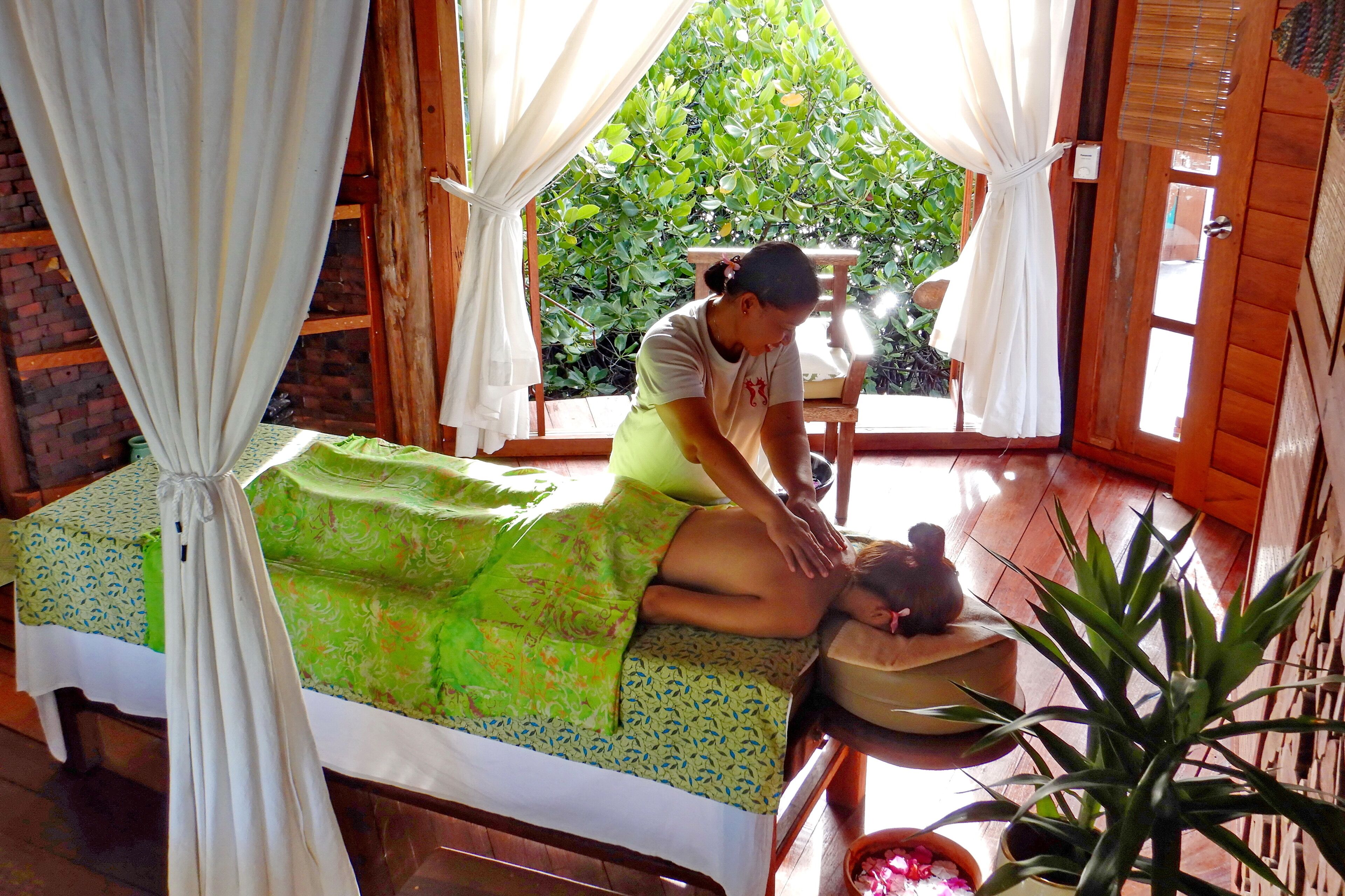 A room with a bed covered by a green patterned bedspread, a wooden table with a bowl, and a potted plant.