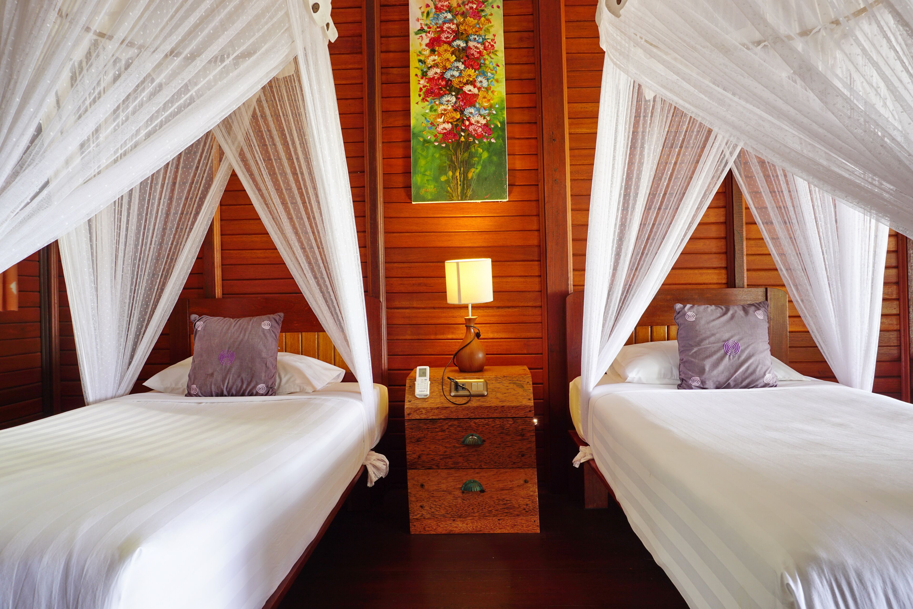A room with two beds, each with white bedding and a canopy. There is a nightstand with a lamp and a vase between the beds.