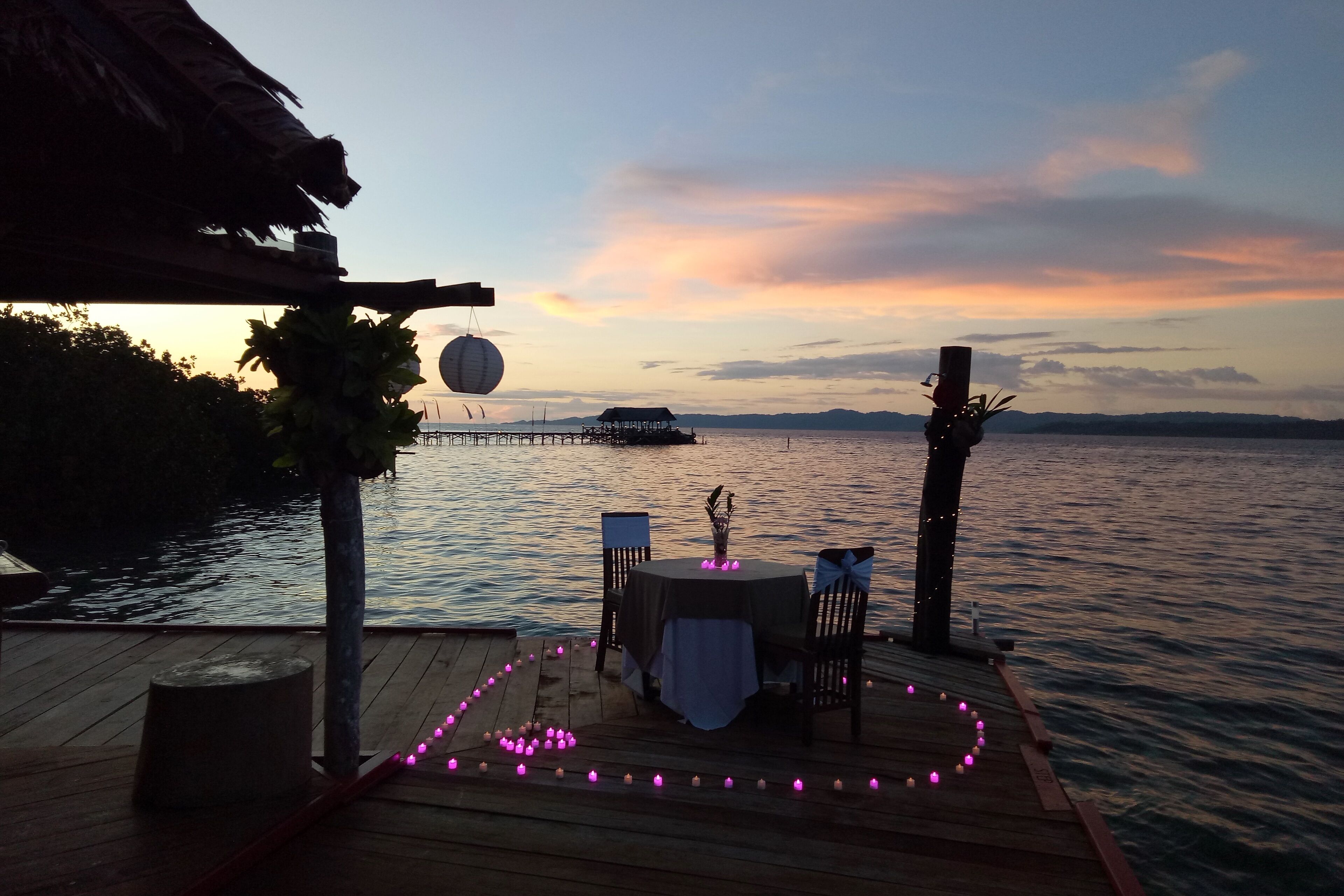 A wooden deck with a table and chairs set up for dining, overlooking a calm body of water with a distant island and a sunset in the background.