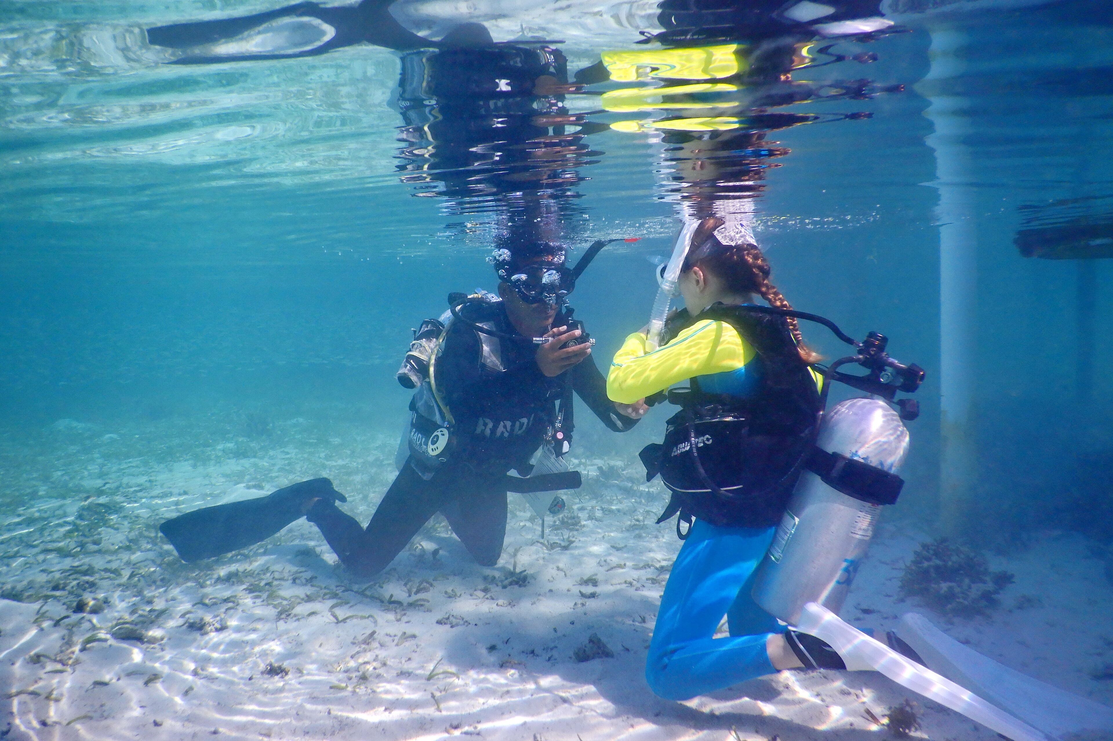Two divers underwater with scuba gear, surrounded by clear water and sandy sea floor.