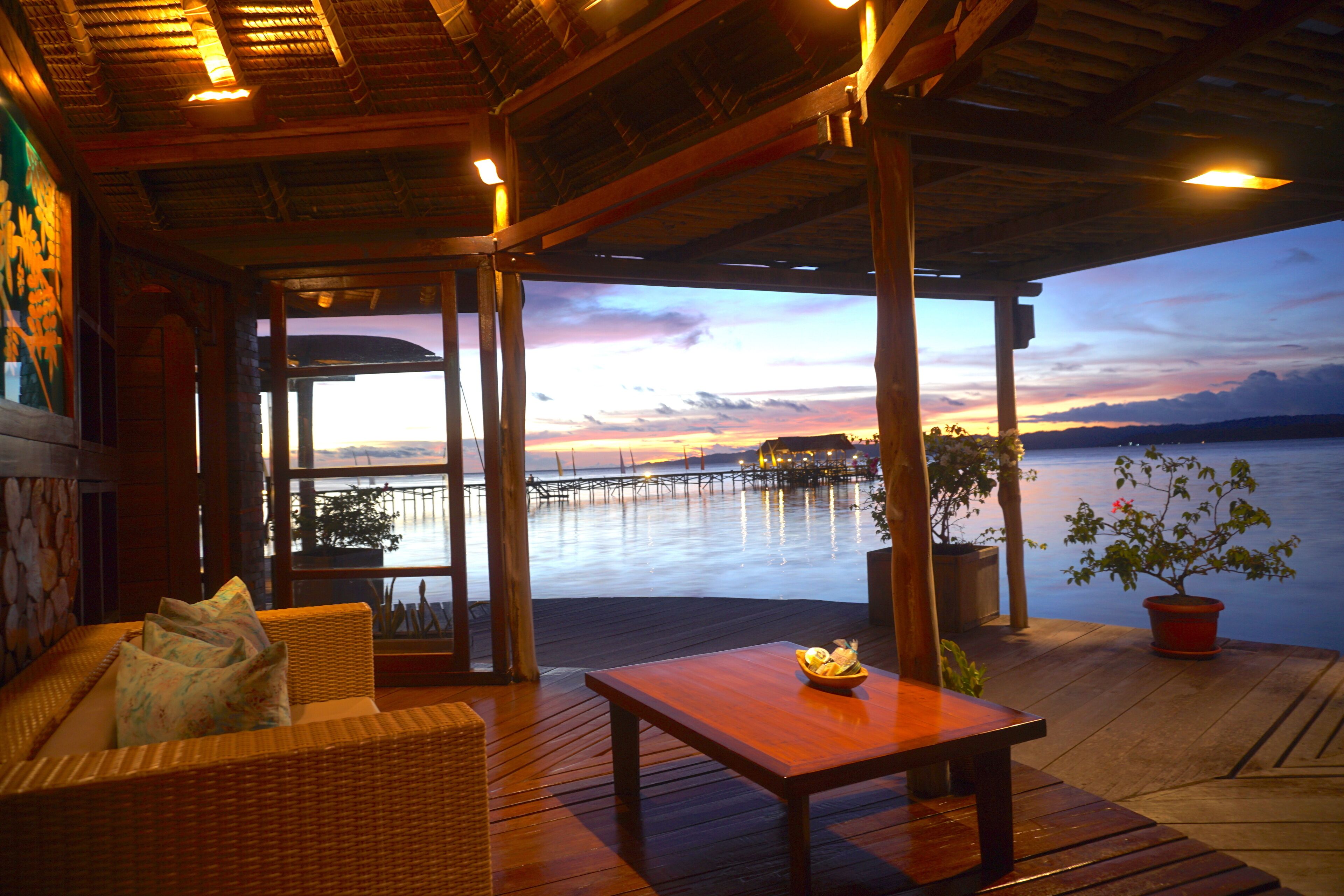 Eine Holzterrasse mit einer Rattan-Couch und einem Tisch, mit Blick auf ein Gewässer und einen Pier im Hintergrund.