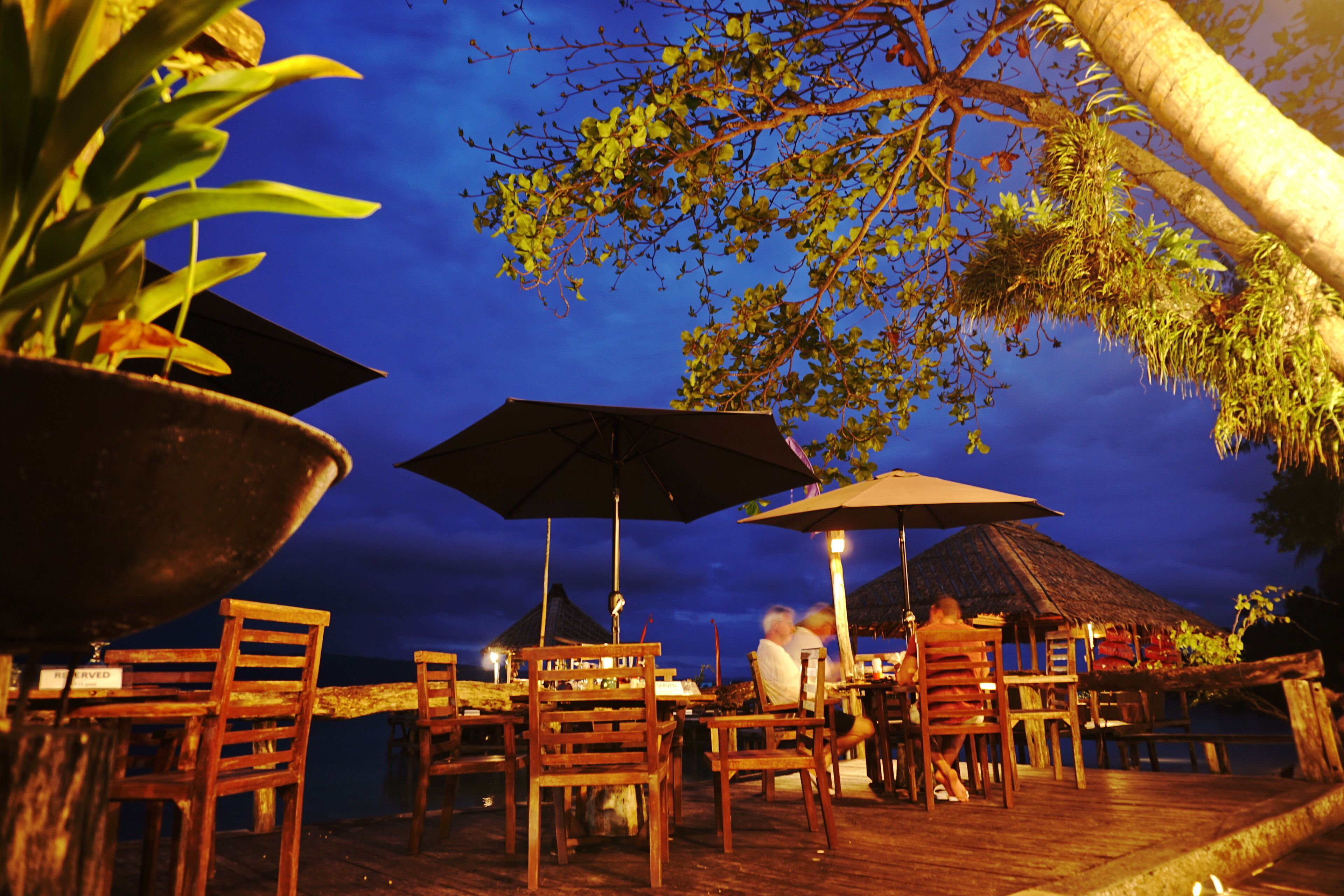 An outdoor dining area with wooden tables and chairs, umbrellas, and a view of the ocean at dusk.