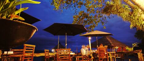 An outdoor dining area with wooden tables and chairs, umbrellas, and a view of the ocean at dusk.