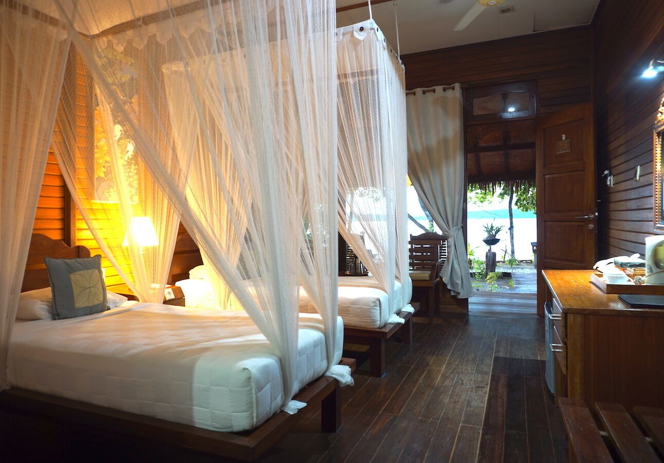 A room with four beds, each with a canopy, wooden flooring, and a small table with a lamp.