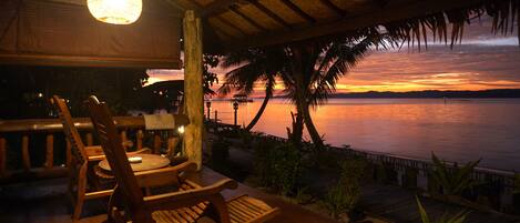 A wooden deck with chairs and a hanging light fixture, overlooking a calm body of water at sunset.