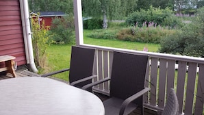 Um alpendre coberto com uma mesa e cadeiras, com vista para um jardim com diversas plantas e um prédio vermelho ao fundo.